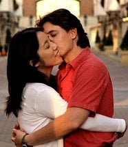 A couple is standing outdoors, embracing each other. The woman has dark hair and is wearing a white shirt, while the man has short dark hair and is wearing a red shirt. They have their eyes closed, and the man is kissing the woman's cheek.