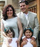 A family of four stands closely together, smiling. The woman has curly dark hair and is wearing a white dress. The man, dressed in a gray suit, stands behind her. In front, two young girls in white dresses, both with headbands, smile brightly.